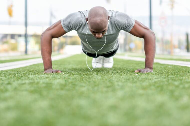 Fitness black man exercising push ups listening to music with headphones. Male model cross-training in urban background. African guy in his twenties doing workout outdoors in the street.